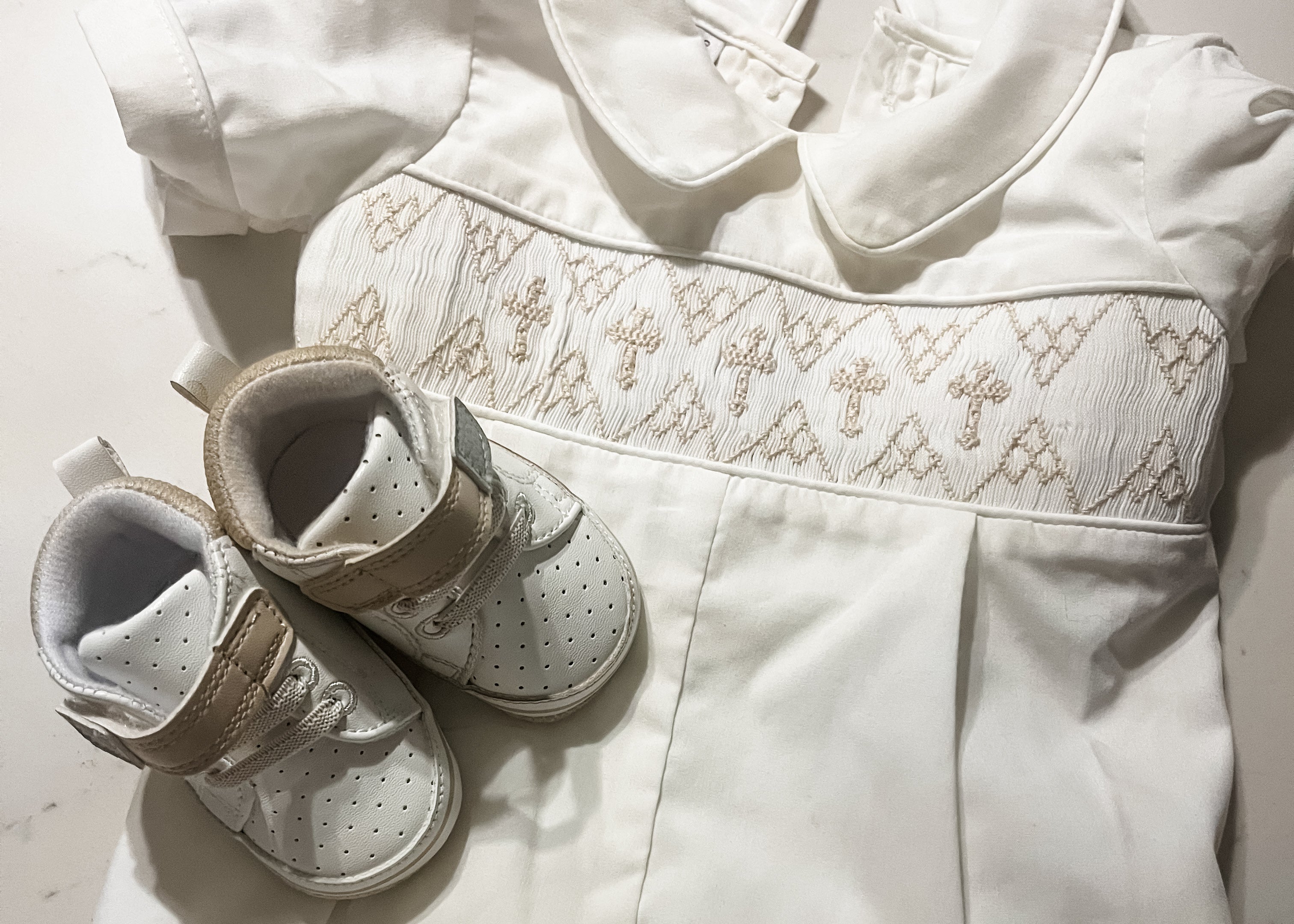 A white baby monstery outfit with a decorative embroidered cross band across the chest, laid flat on a light marble surface alongside a pair of white and beige baby shoes.