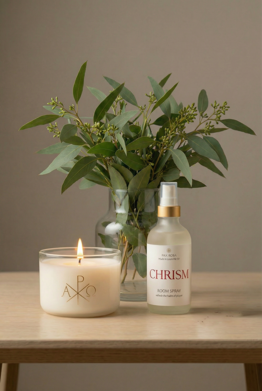 a candle with the Chi Rho image and room spray bottle with label "Chrism" on a table with a vase of greens