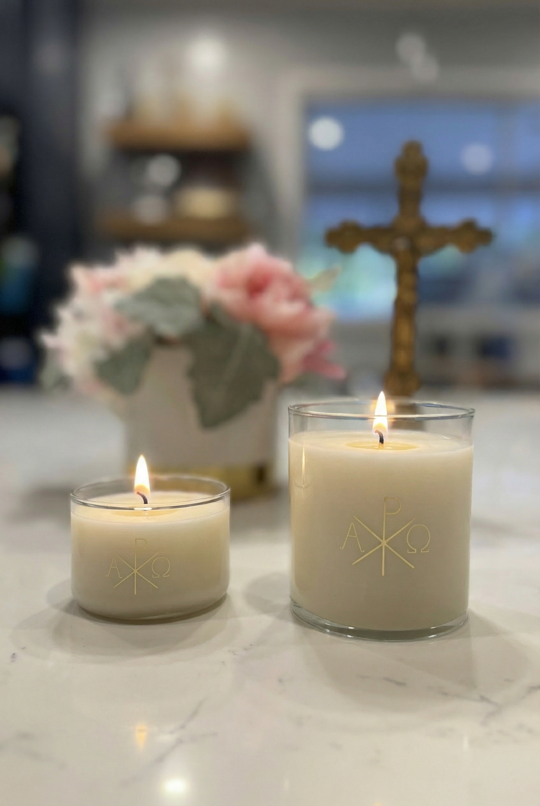 Two lit candles on a marble surface with a decorative cross and flowers in the background.